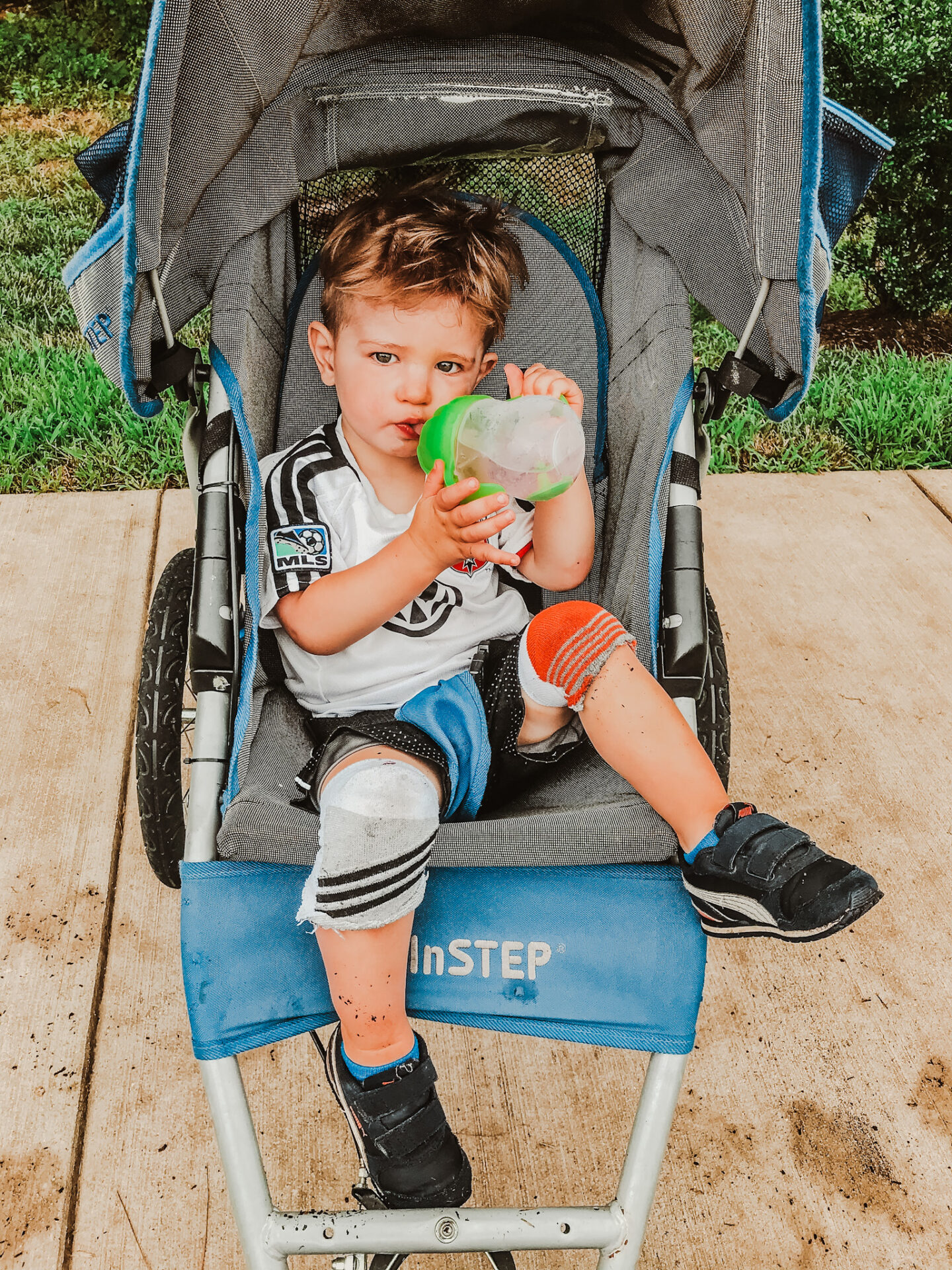Toddler boy sitting in blue stroller drinking water wearing sock knee pads.