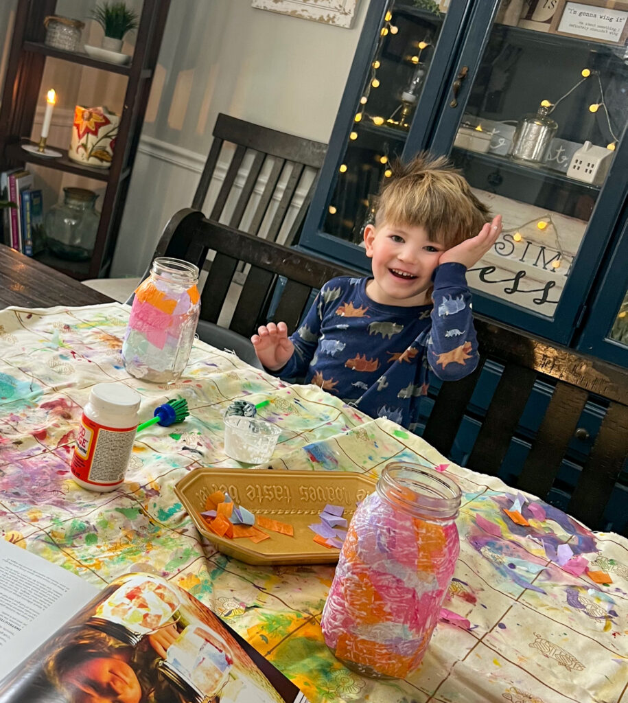 Young boy crafting a mason jar lantern with Mod Podge, tissue paper, and a mason jar at the dinning room table.