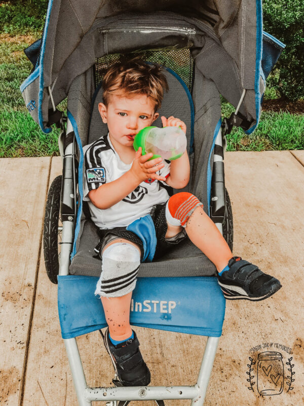 Toddler boy sitting in blue stroller drinking water wearing sock knee pads.