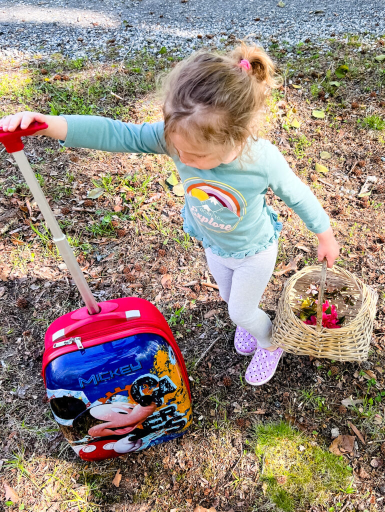 Young girl holding a basket in one hand and a rolling suitcase in the other for a scavenger hunt in nature.