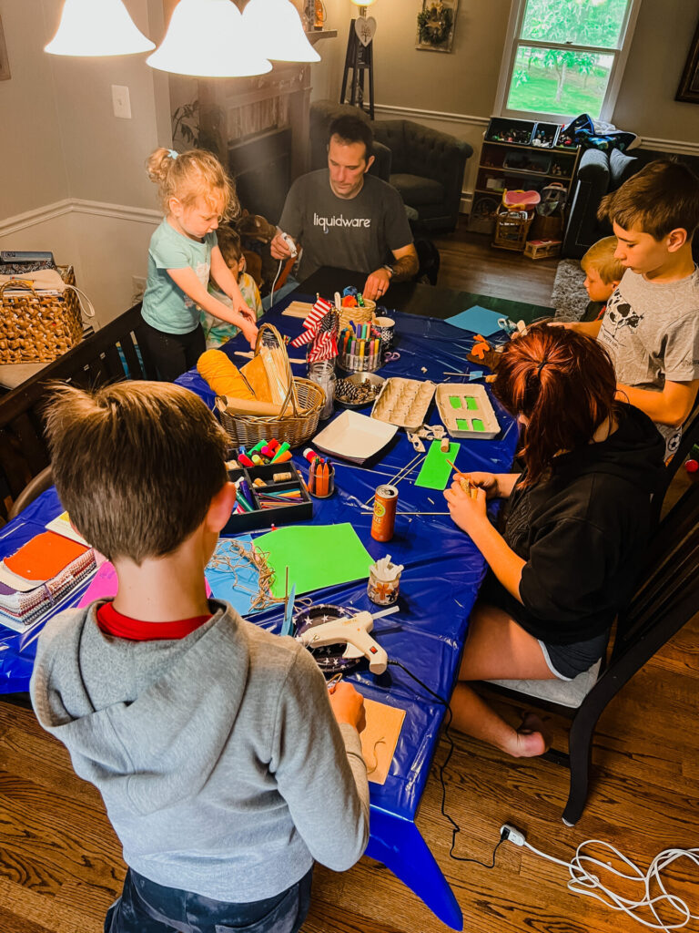 Table view of people of all ages working on DIY boats with craft materials scattered on the table.