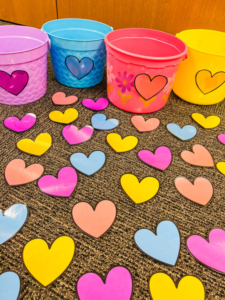 Color Matching Game for Valentine's Day. Perfect for Toddlers and Preschool children. 4 plastic buckets, pink, yellow, blue, and purple. In front of the buckets are an assortment of pink, yellow, blue, and purple hearts.